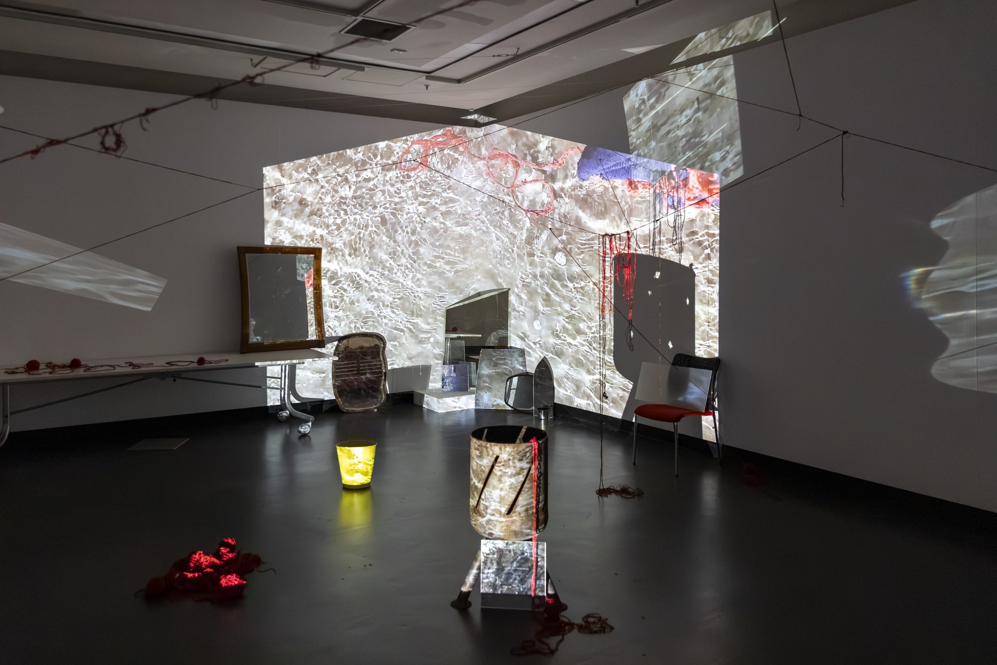 Projections on the walls, a mirror and lit containers among red yarn on the gallery floor