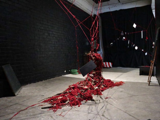 Red yarn descending from the ceiling into a heap on a gallery floor in low light